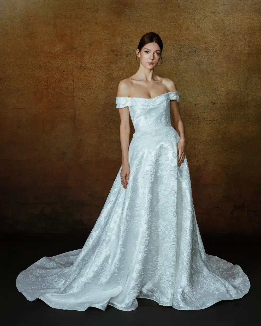 A woman in an elegant off-the-shoulder white gown with a full skirt, standing against a textured brown background.