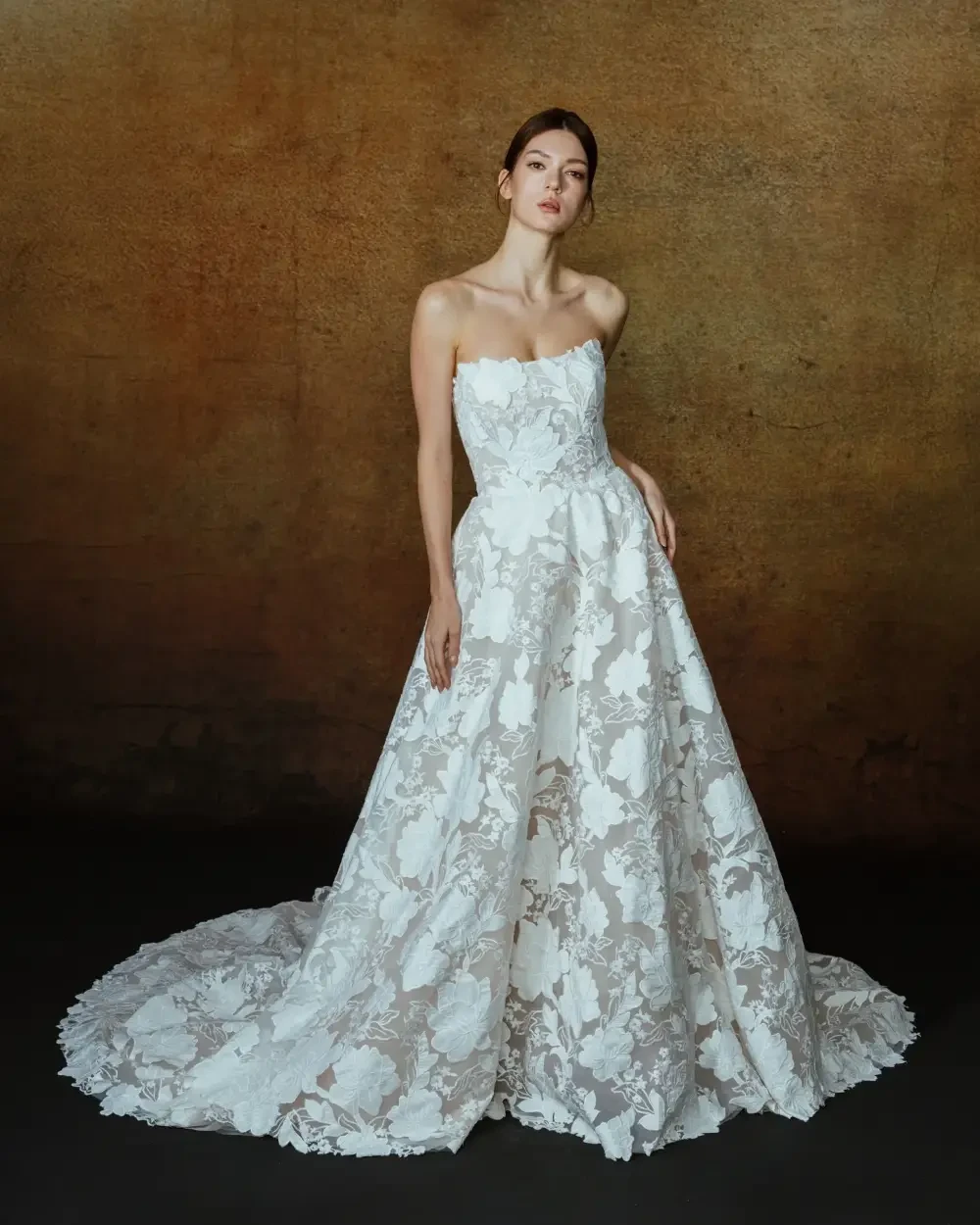 A model wearing a strapless, white wedding gown with intricate lace patterns and a flowing train, posed against a textured brown backdrop.